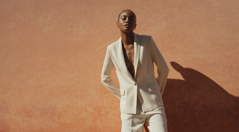 A model standing in front of a wall wearing a white single breasted blazer with nothing underneath. The blazer is matched with a pair of white wide leg trousers.