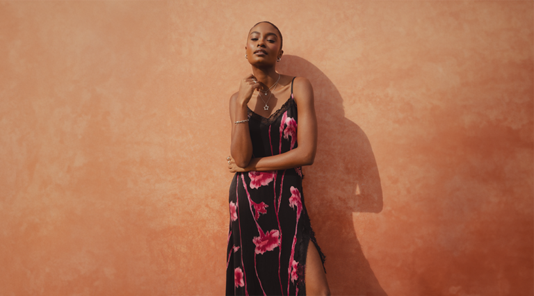 A model standing in front of a clay wall wearing the Saint and Sofia Harper Lace Cami Dress in Pink Stem Floral. This floral dress features a black background with bright pink flowers. It has a lace hem and cami-style straps.