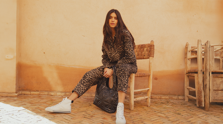 A model sat on a rattan chair wearing a leopard print denim jacket from Saint and Sofia with a pair of matching leopard print jeans. She styles them here with white high top trainers and a black quilted nylon bag.