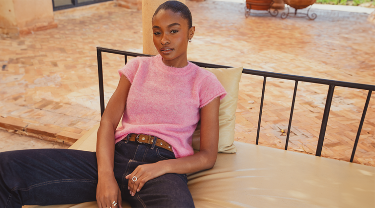 A model sat on an outdoor sofa wearing the Saint and Sofia Ariella Baby Tee in pink with a pair of the Indigo barrel leg jeans.