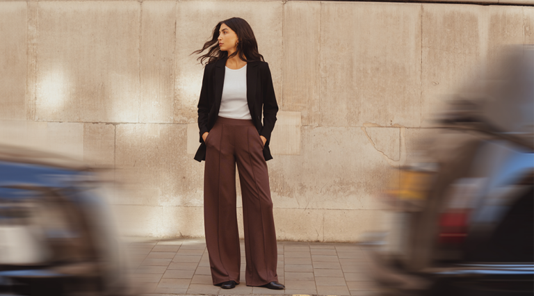 A model standing on a London street in front of a brick building. She in front of her on the road two taxi's whizz past in the street. She is framed between the two cars. She looks off into the distance wearing a white slim-fit tee by Saint and Sofia with a black double-breasted blazer and brown wide leg trousers with a front pin tuck detail down each leg.