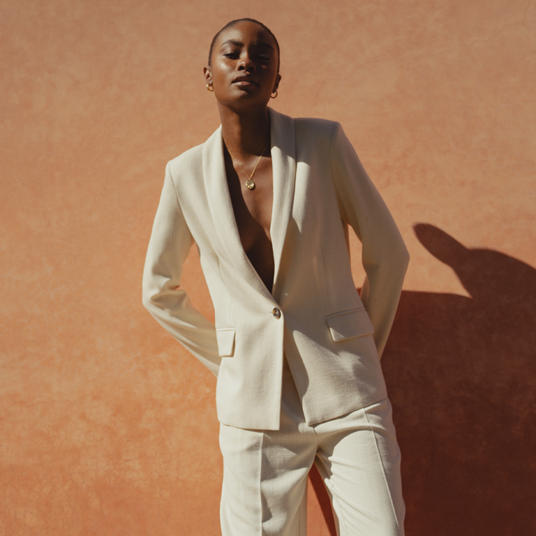 A model standing in front of a wall wearing a white single breasted blazer with nothing underneath. The blazer is matched with a pair of white wide leg trousers.