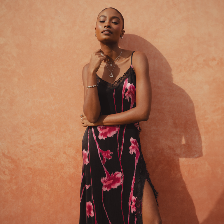 A model standing in front of a clay wall wearing the Saint and Sofia Harper Lace Cami Dress in Pink Stem Floral. This floral dress features a black background with bright pink flowers. It has a lace hem and cami-style straps.