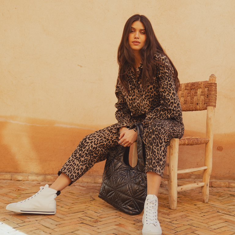 A model sat on a rattan chair wearing a leopard print denim jacket from Saint and Sofia with a pair of matching leopard print jeans. She styles them here with white high top trainers and a black quilted nylon bag.