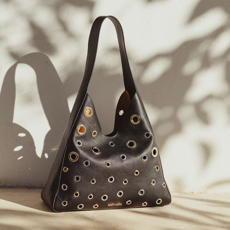 The Allana Eyelet Bag by saint and sofia. A leather hobo bag style with silver and gold metal eyelet details across the front. It's photographed here in front of a wall with the shadows of a tree scattered across the wall behind it.