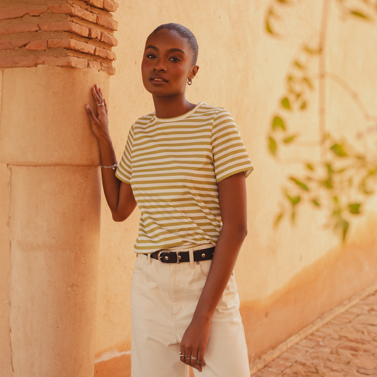 A model leaning against a stone pillar wearing a green and white striped crew neck, short sleeve T-shirt. Styled here tucked into a pair of white jeans with a black leather belt.