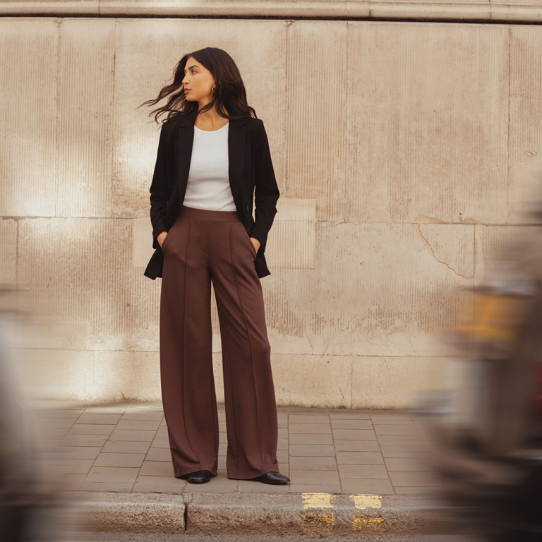 A model standing on a London street in front of a brick building. She in front of her on the road two taxi's whizz past in the street. She is framed between the two cars. She looks off into the distance wearing a white slim-fit tee by Saint and Sofia with a black double-breasted blazer and brown wide leg trousers with a front pin tuck detail down each leg.