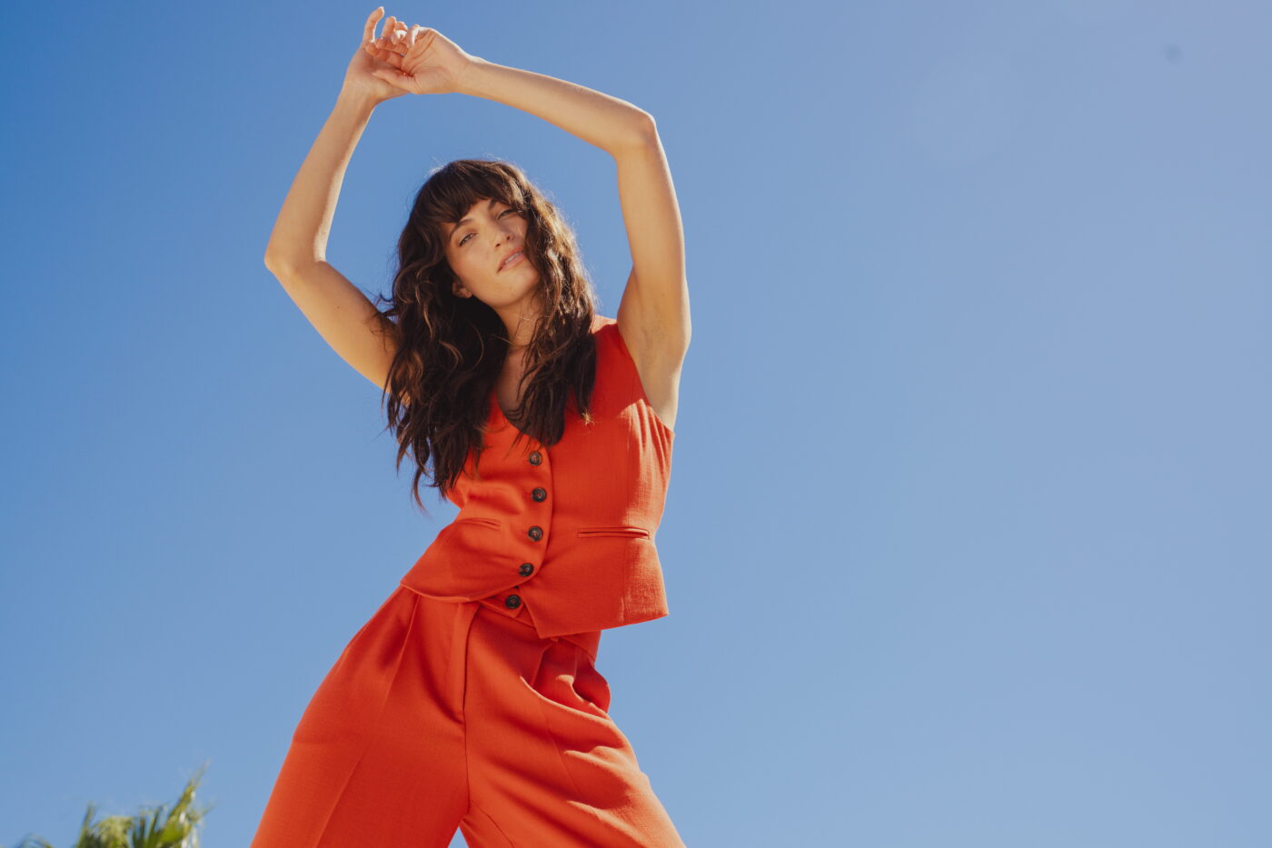 An image of a model under a blue sky with her hands above her head wearing the Burnt Orange waistcoat Astrid Waistcoat by Saint and Sofia with the matching Argent Wide Leg Trousers