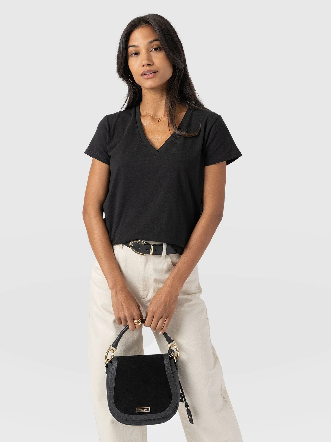 A model wearing the Apartment Tee by Saint and Sofia. A relaxed, V-neck style with short sleeves. Worn here tucked into a pair of white jeans with a black leather belt and black leather saddle bag.