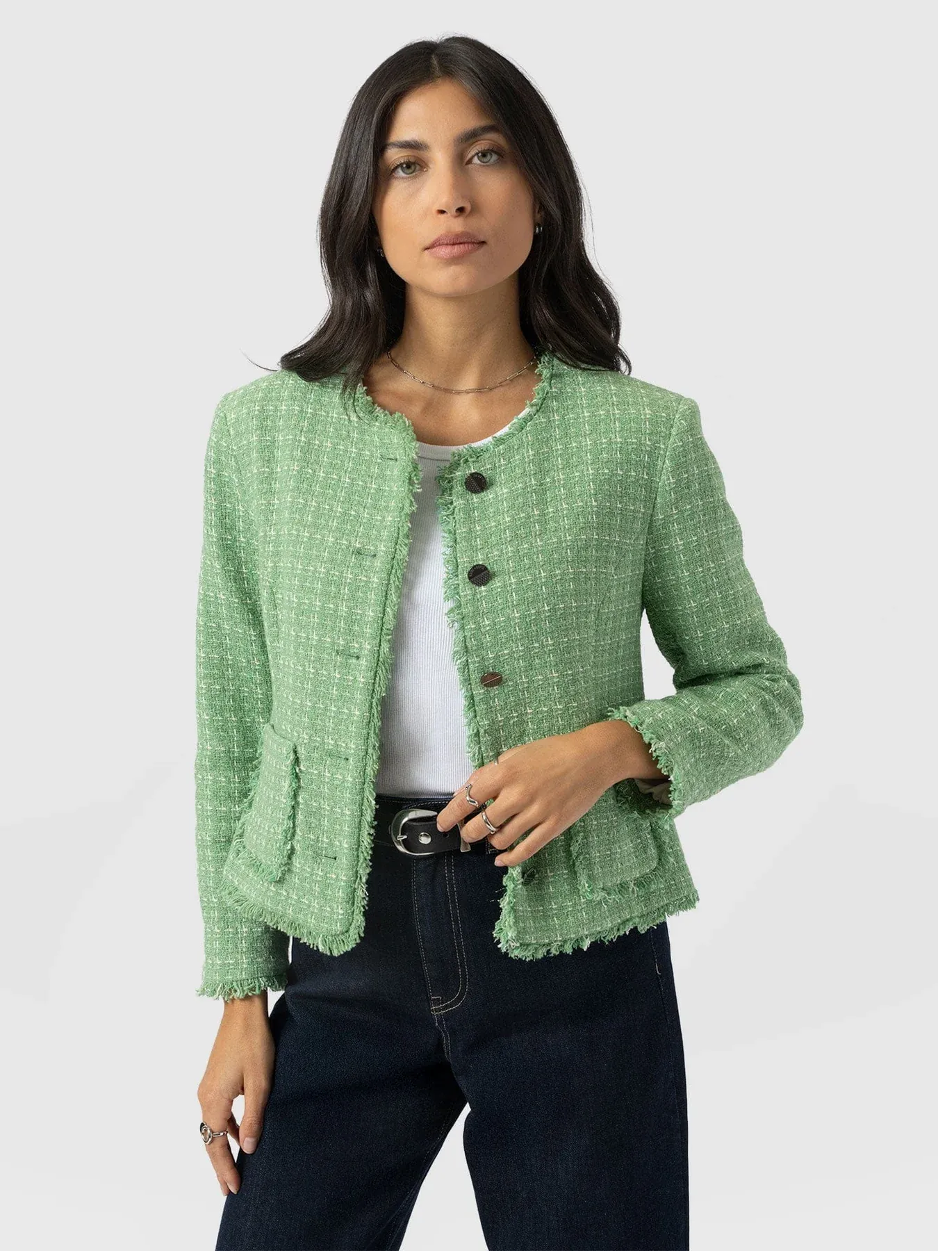 A model wearing the Ashby Jacket by Saint and Sofia, a boucle design in a mint colour with frayed detailing along the trims. Worn here over a white tank top and dark denim jeans.