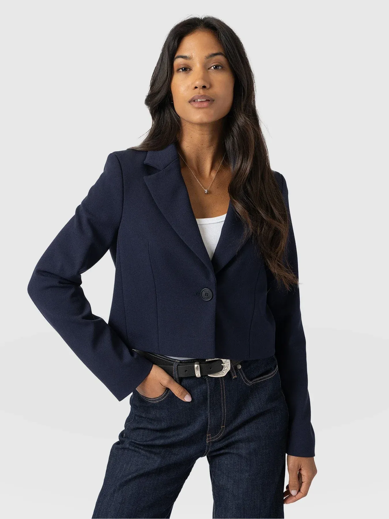 A model wearing the Saint and Sofia Astrid Cropped Blazer in Navy. A Single breasted design worn over a white tank top and indigo denim jeans.