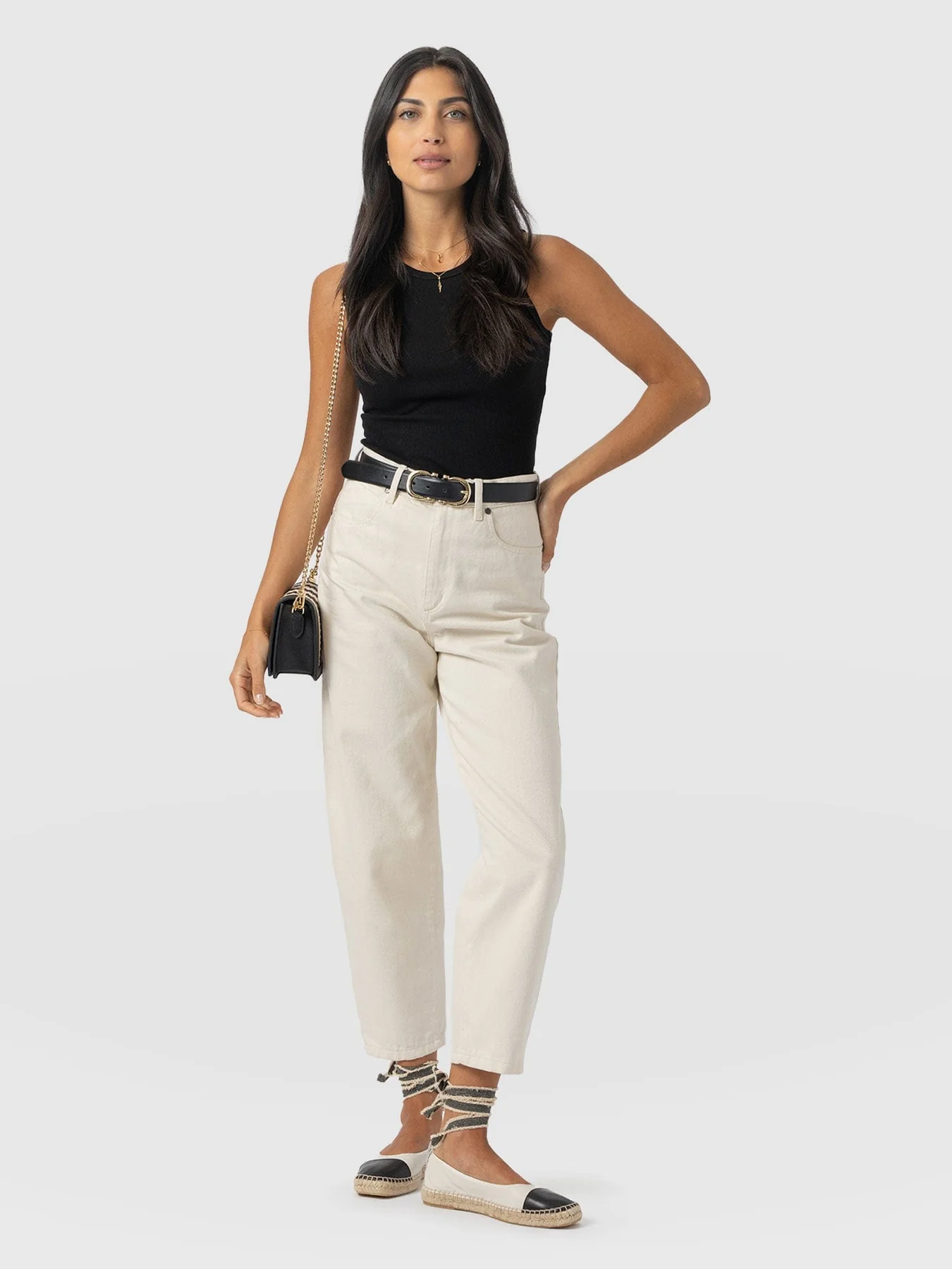 A model wearing a pair of cream barrel leg jeans with a black sleeveless vest tucked in. They're styled here with a black leather belt and wrap-around espadrille flats with a leather toe detail.