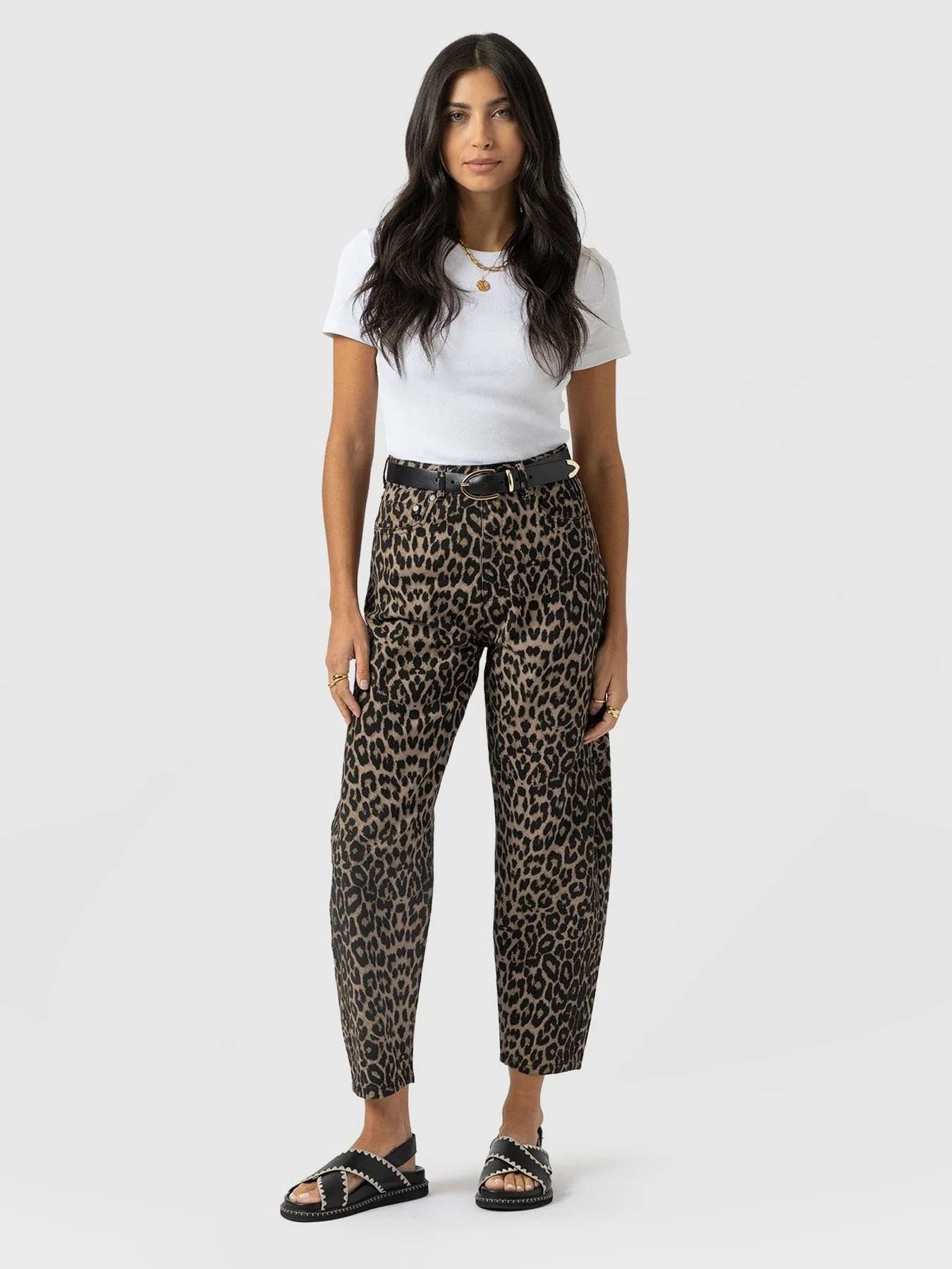 A model wearing a pair of leopard print barrel leg jeans with a white short sleeve top, leather belt, and suede cross-over sliders.
