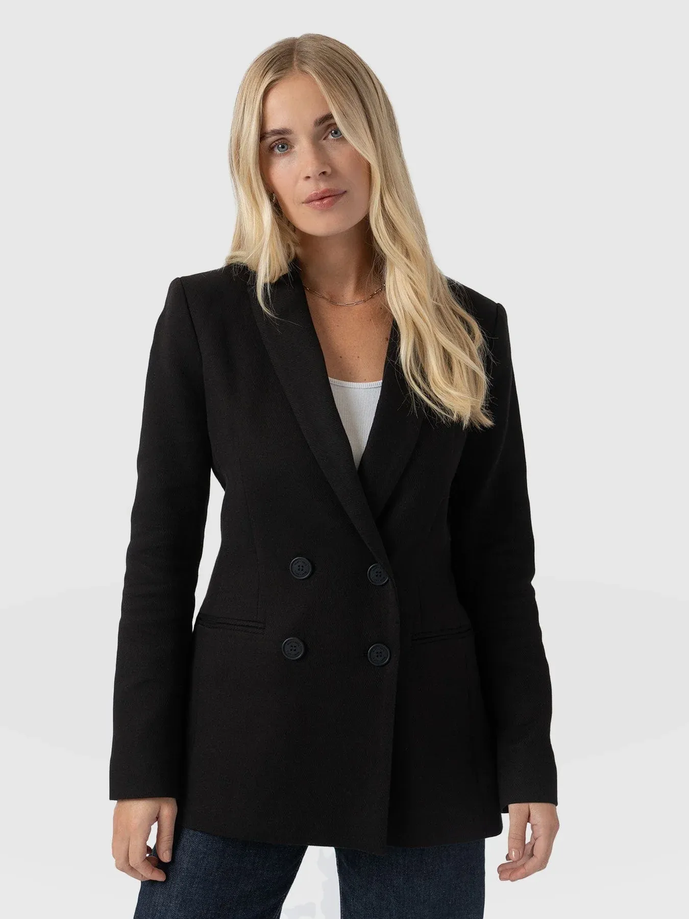 A model wearing the Cambridge Blazer by Saint and Sofia. A double breasted black blazer with a classic notched lapel collar. Worn here over a white tank top with denim jeans.