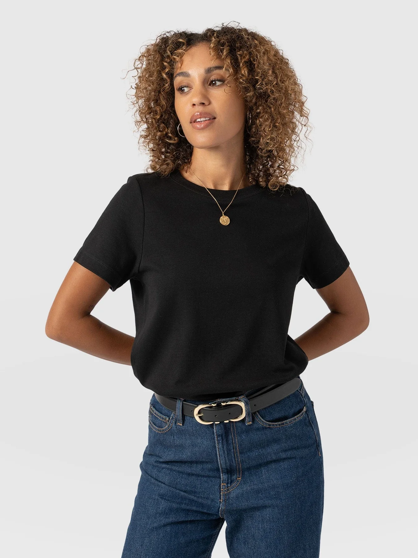 A model wearing the black Chelsea Crew Neck T Shirt by Saint and Sofia. A boxy crew neck style with short sleeves. It's styled here tucked into a pair of denim jeans with a black leather belt and a cold pendant necklace.