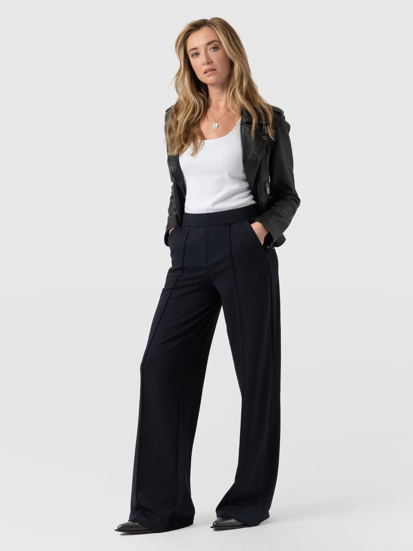 A model standing in front of a white back drop wearing the Saint and Sofia Keller Pant. A wide leg tailored-effect trouser with a flat waistband and front pintuck detail. Styled here with a white scoop neck vest and a black leather jacket.