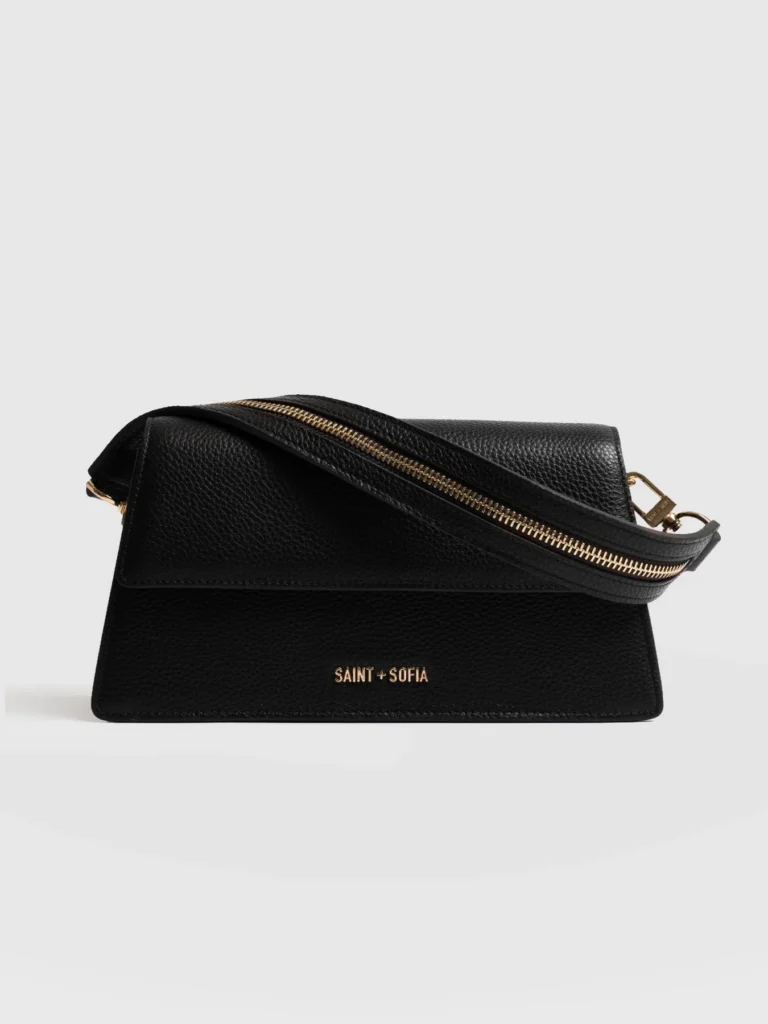 A black leather bag from Saint and Sofia, sculpted to a trapeze shape with a metal branded logo at the front and an adjustable canvas shoulder strap