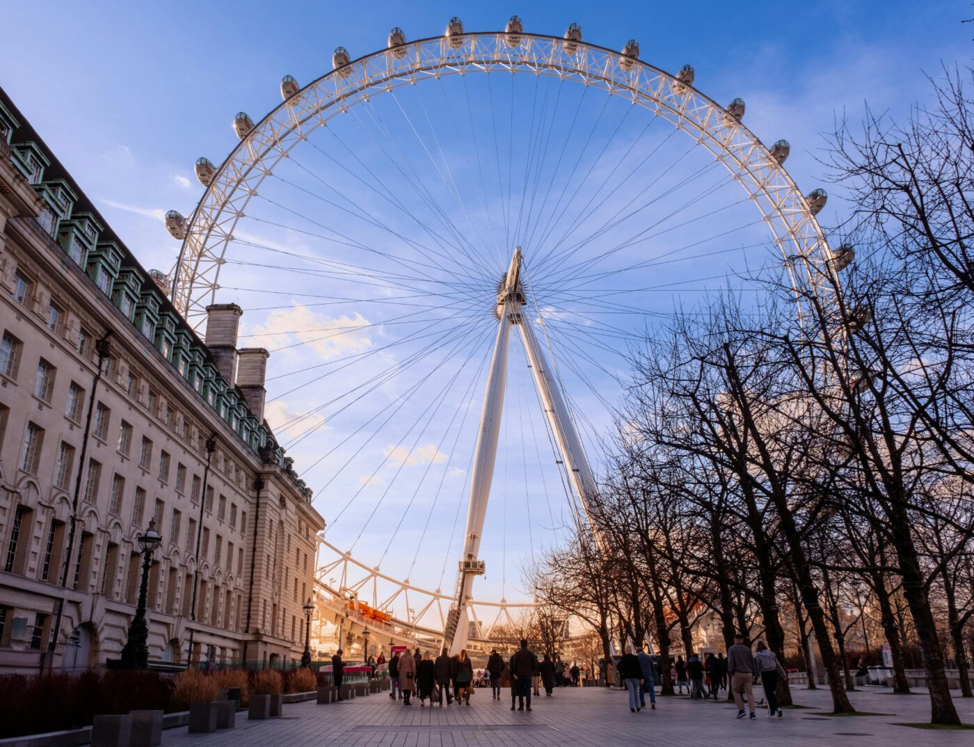 A low down picture of the London Eye taken at sunset. At the bottom of the frame, tourists walk down the street.