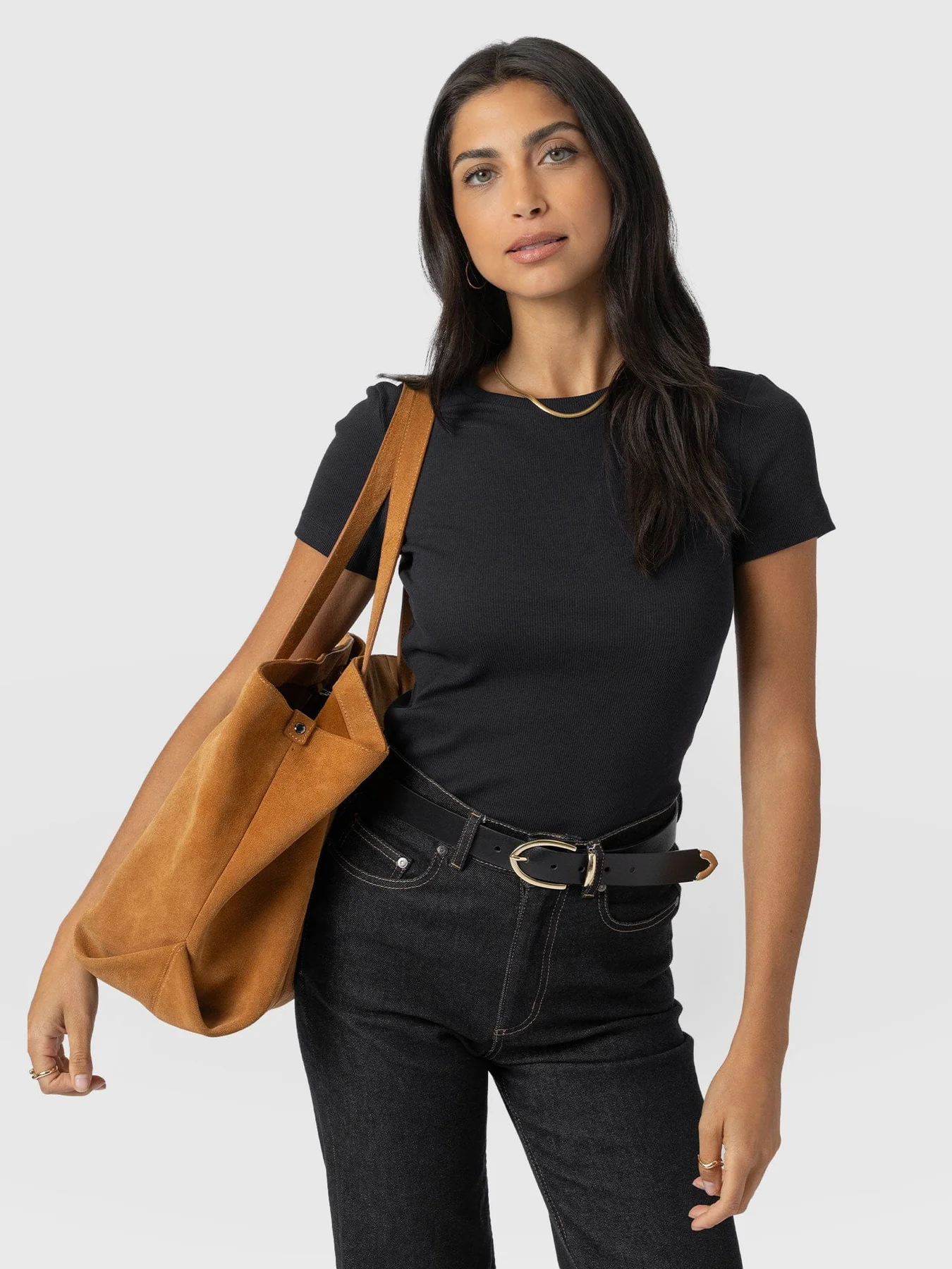 A model wearing the Saint and Sofia Ribbed Short Sleeve T Shirt. A crew neck style with a slim fit and short sleeves. Here, it's styled with black jeans, a leather belt, a gold chain necklace, and a brown suede tote bag.