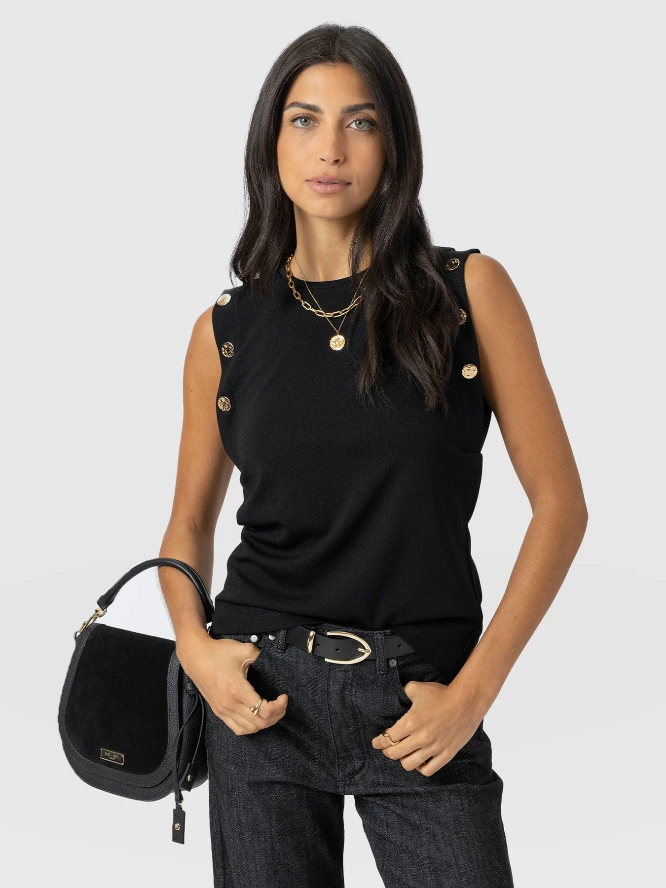 A model wearing the Rowen Tee from Saint and Sofia. A crew neck, sleeveless style with gold molten button details at the arms. It's worn here tucked into a pair of black jeans with a black leather belt and black leather saddle bag.