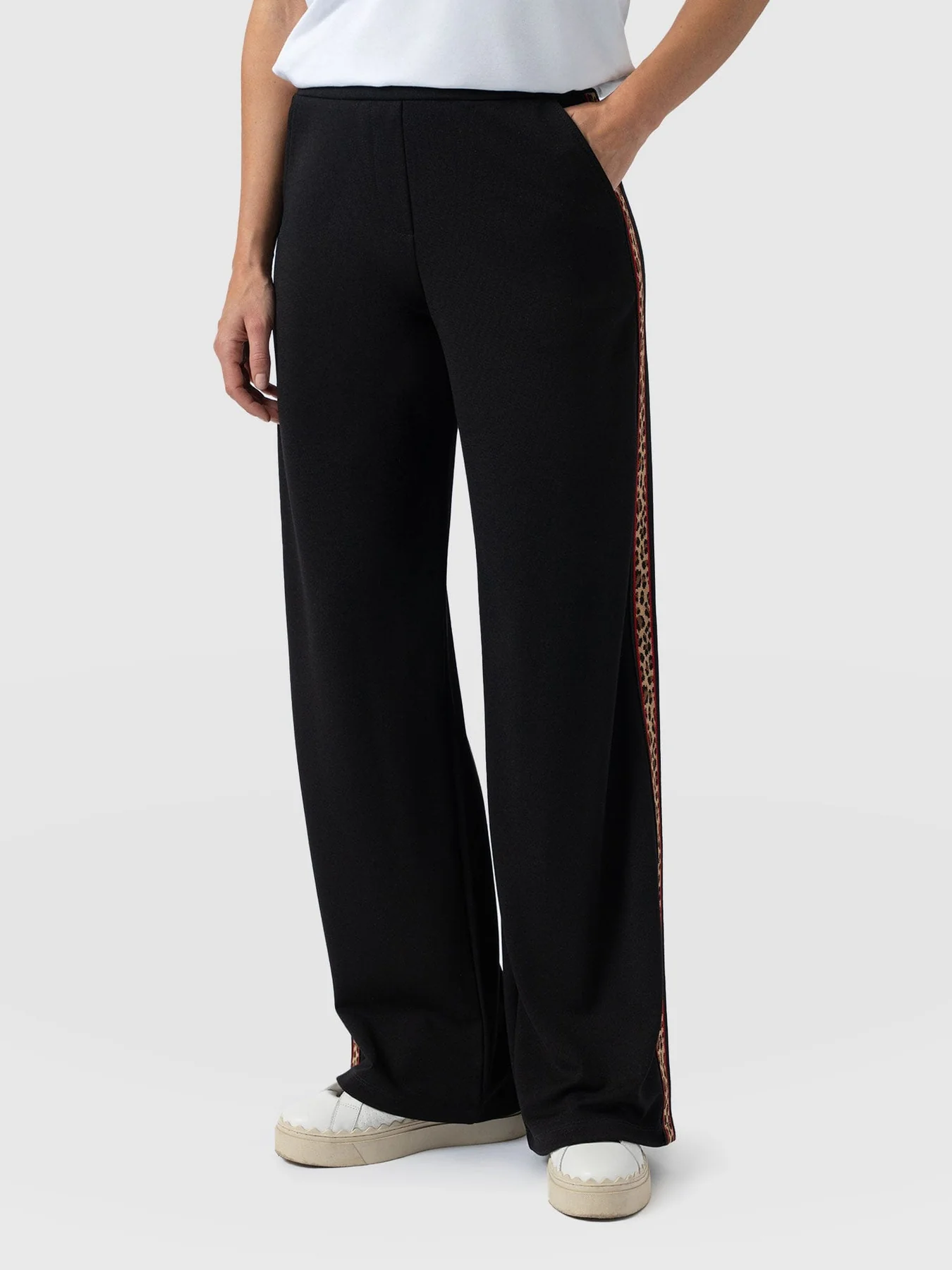 A close up shot of the Victory Pant by Saint and Sofia, wide leg trousers in black made from a soft stretch jersey with leopard print taping down the leg. Styled here with white leather trainers.