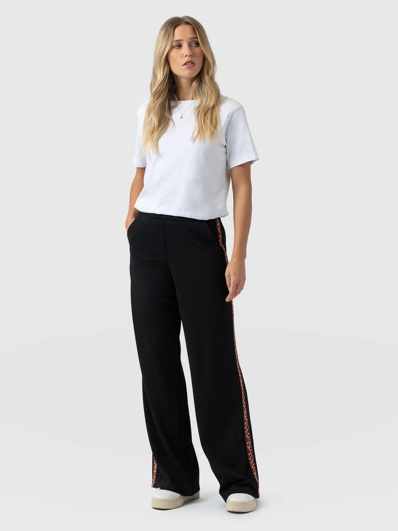 A model stood in front of a white backdrop wearing the Saint and Sofia Victory Pant, a wide leg loungewear trouser with contrasting leopard taping down the leg. Worn here with a white crew neck short sleeve tee and white trainers.