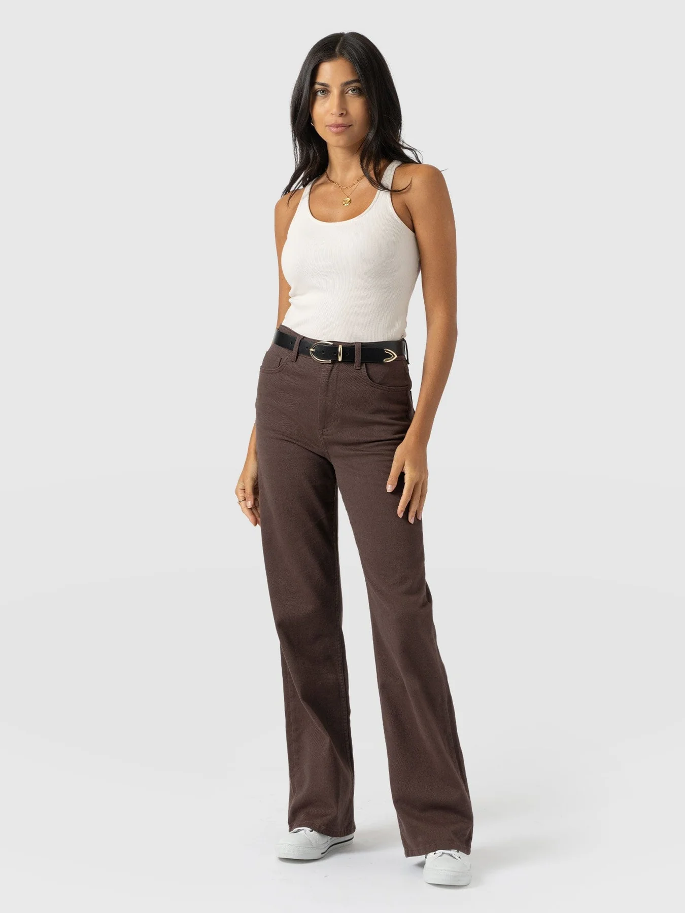 A model wearing brown wide leg jeans with a white tank top tucked into the high rise waistband. She wears it with a black leather belt and white leather trainers.