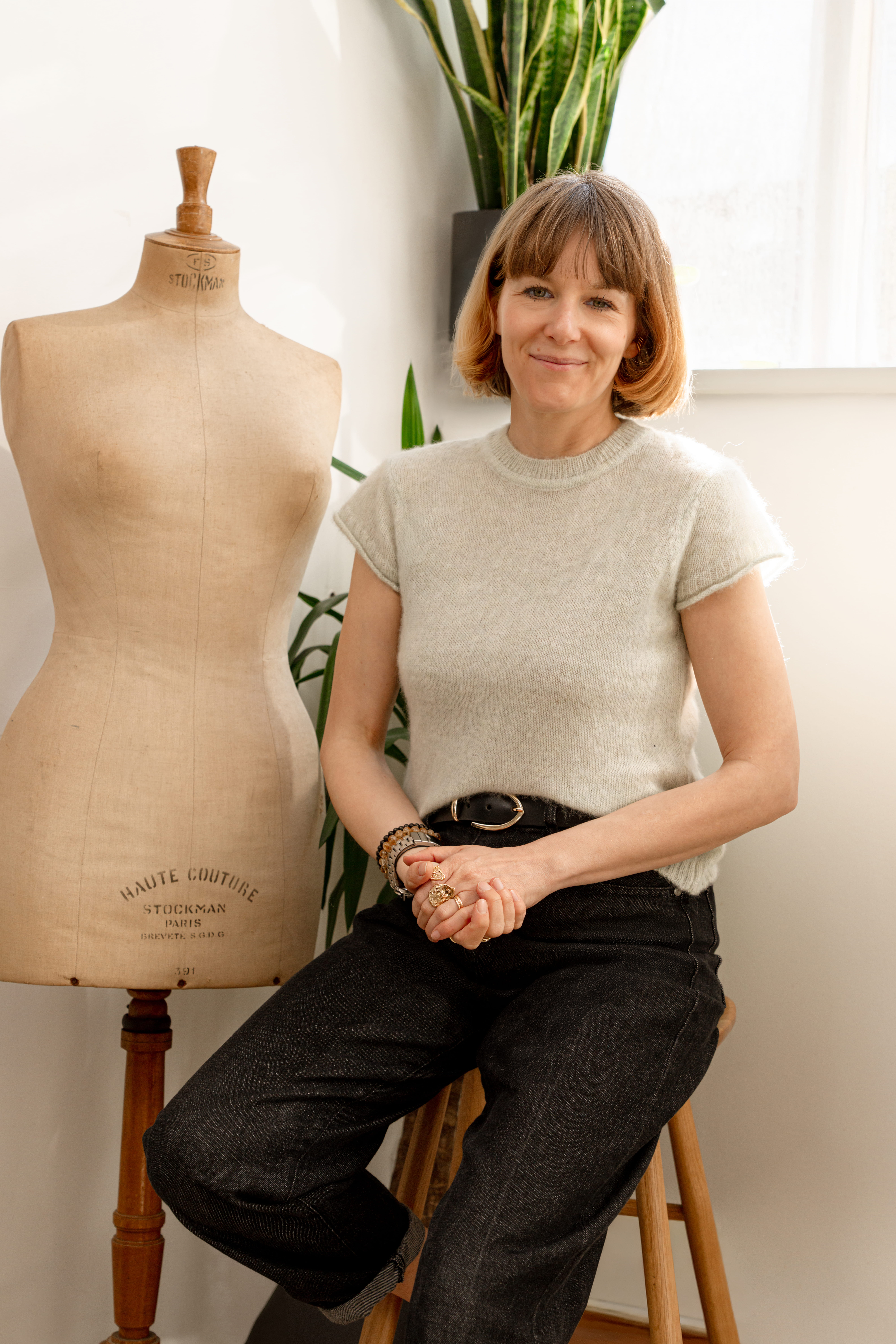 An image of the Head of Editorial at Saint and Sofia, Alexandra Fullerton. She sits on a stool in the Saint and Sofia studio wearing the Ariella Baby Tee in beige and a pair of black jeans. She sits beside a mannequin torso and in front of a plant. Her hands are clasped across her lap.