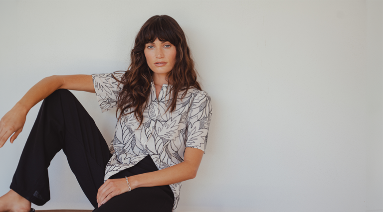 A model sitting on a wooden bench against a white wall. She wears a white short sleeved blouse with black floral embroidery and a pair of wide-leg black linen trousers. She sits with one knee bent with her foot on the bench and her arm resting over the knee.