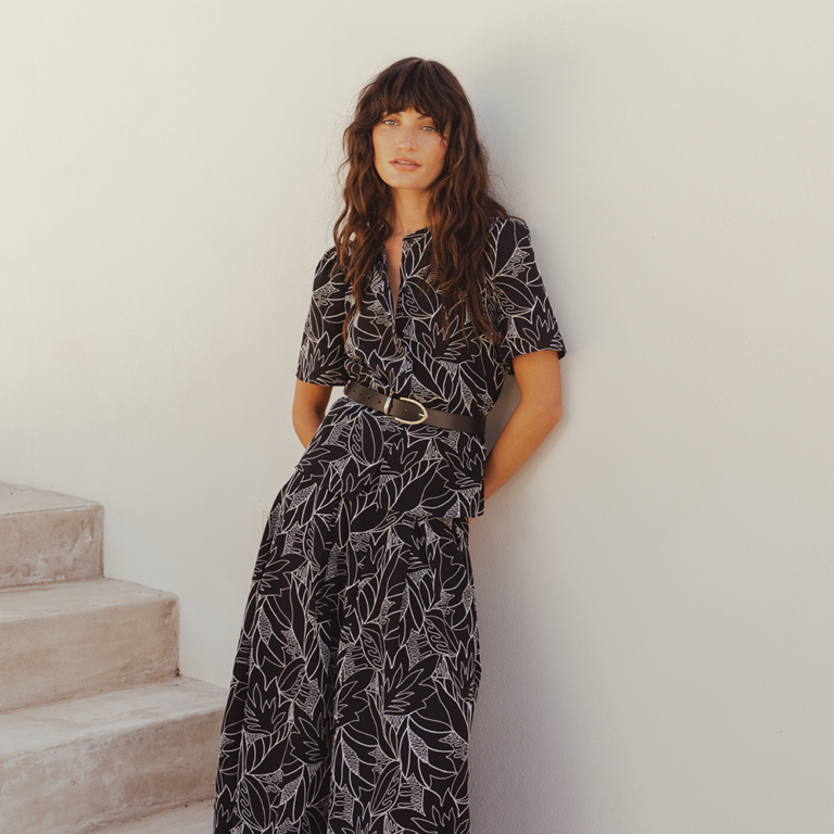 A model leaning against a white clay wall beside a stone staircase. She wears the Saint and Sofia Tabitha Blouse and Eliza Skirt in Black Embroidered. Black cotton styles with contrasting white floral embroidery. She styles the shirt with a belt at the waist to make the two-piece mimic a dress.