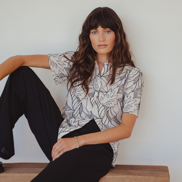A model sitting on a wooden bench against a white wall. She wears a white short sleeved blouse with black floral embroidery and a pair of wide-leg black linen trousers. She sits with one knee bent with her foot on the bench and her arm resting over the knee.