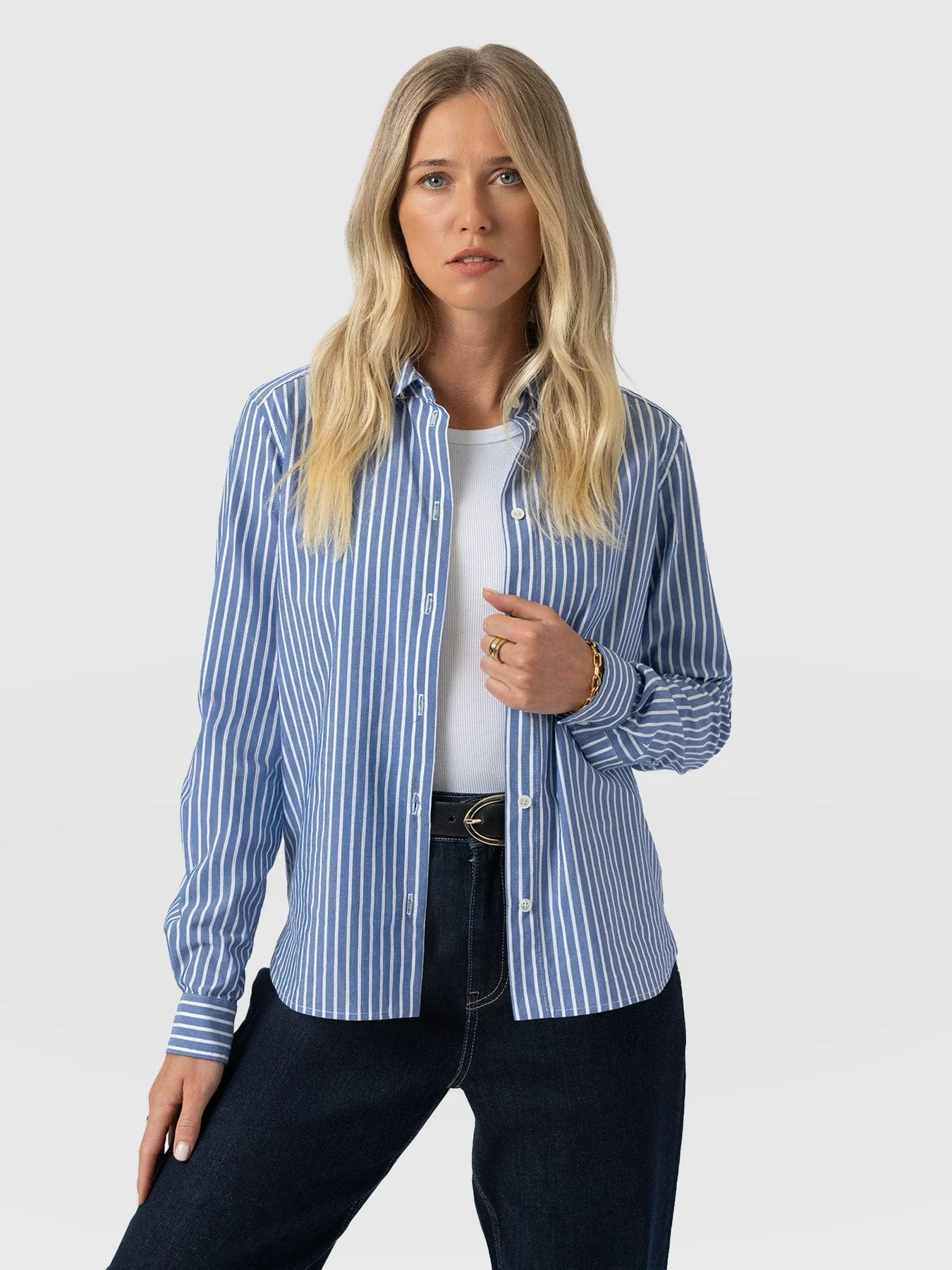 A model wearing the Anneliese Shirt by Saint and Sofia in blue and white stripe. She wears it unbuttoned over a white ribbed cami and over a pair of indigo denim jeans with a belt at the waist.