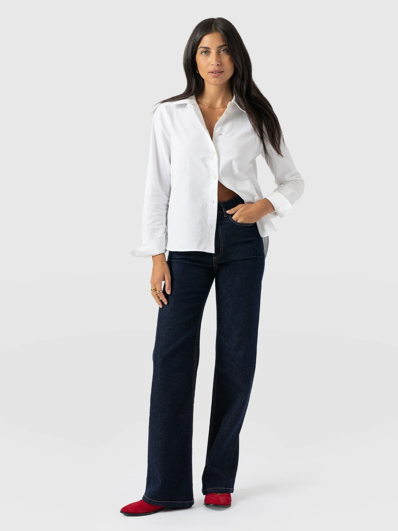 A model wearing the Anneliese Shirt by Saint and Sofia, a classic white button up shirt made from Oxford Cotton Poplin. She wears it here open at the hem with a pair of indigo wide leg jeans and red ballerina flats.