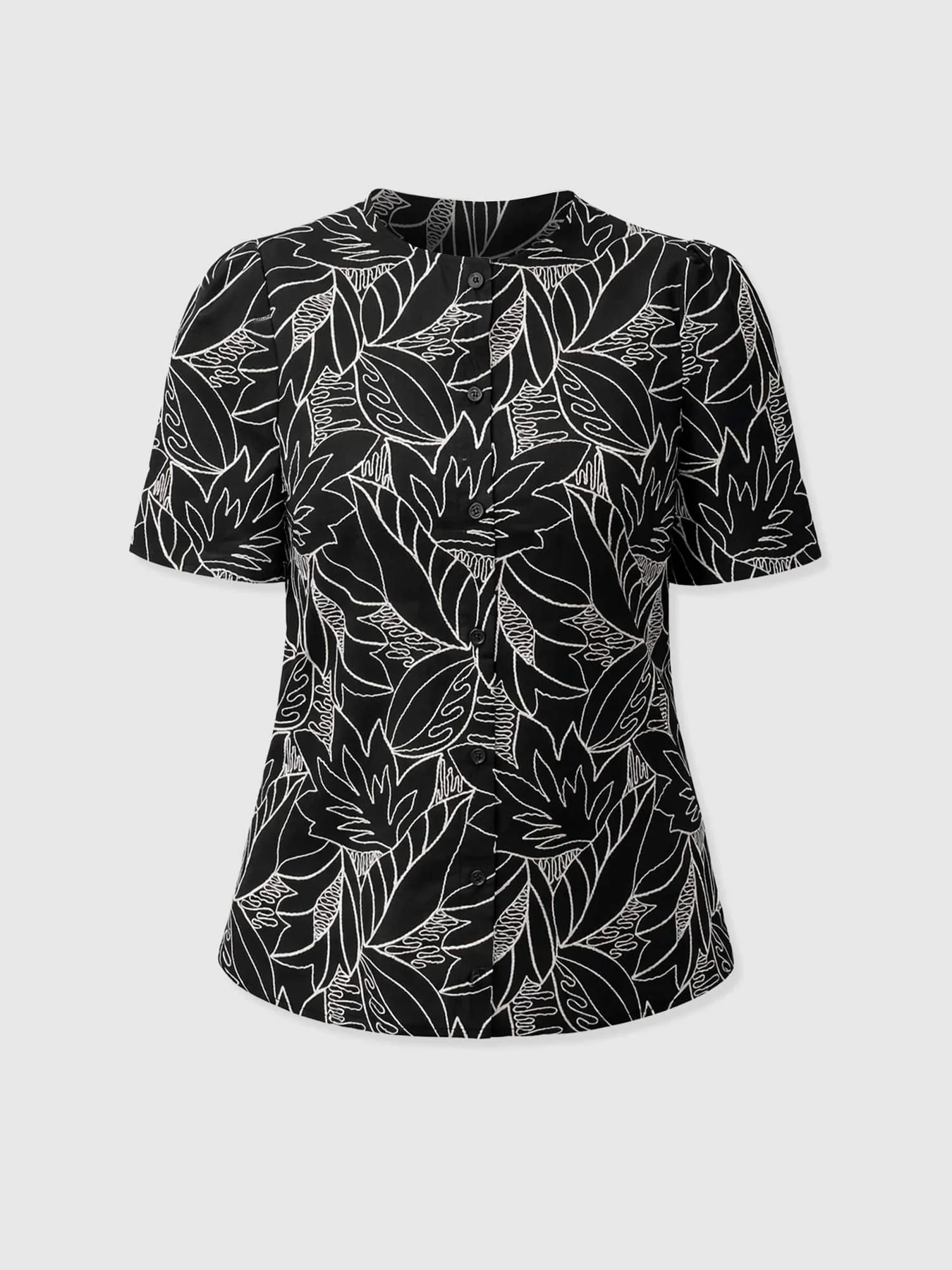 A cut out image of the Tabitha Short Sleeve Blouse in Black Embroidered by Saint and Sofia. This cotton style features a button-up fastening with lightly puffed sleeves, a black background, and contrasting white stitching.