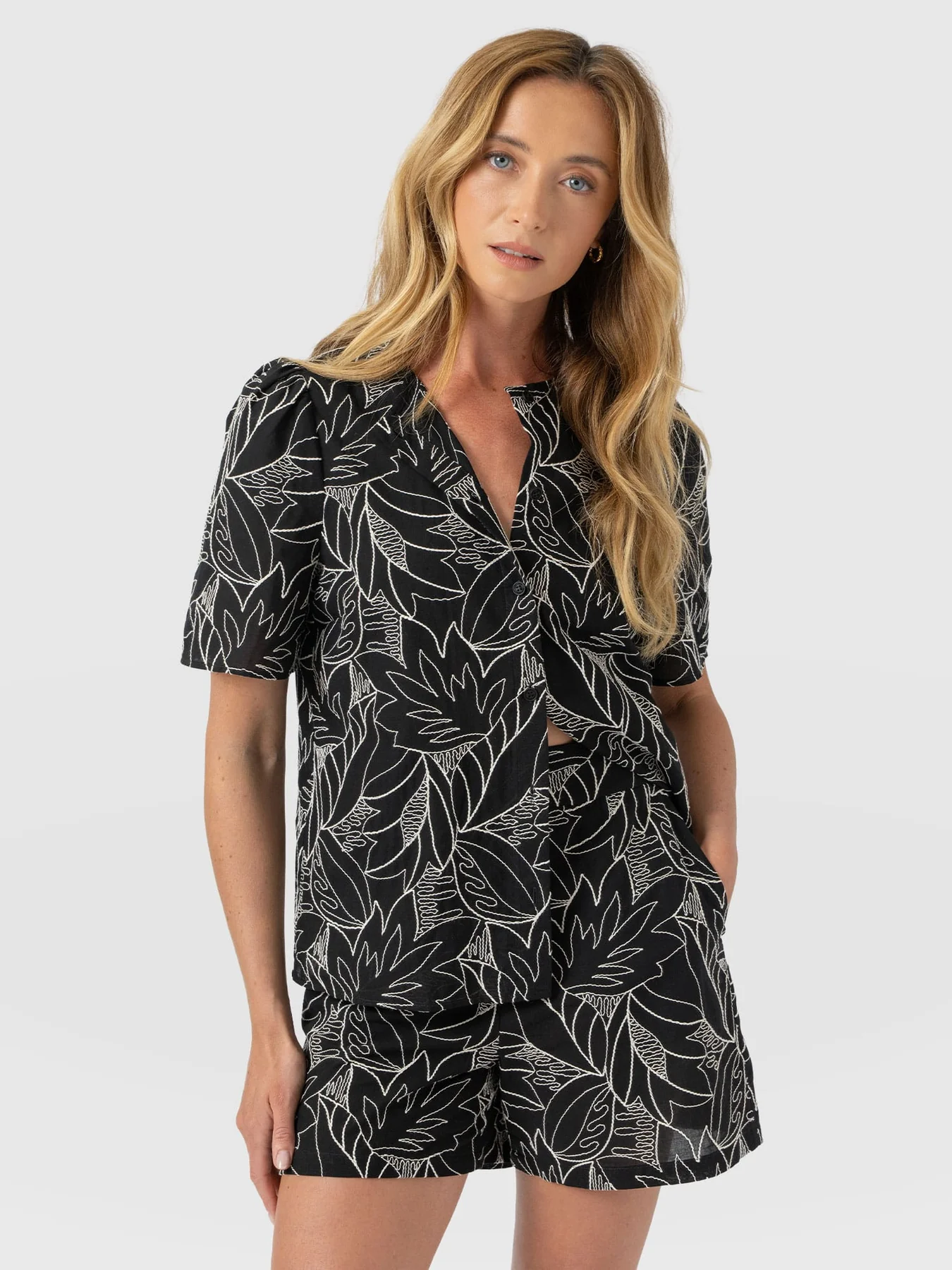 A model standing in front of a white background. She wears the Tabitha Short Sleeve Blouse in Black Embroidered by Saint and Sofia and a pair of the matching shorts. This cotton style features a button-up fastening with lightly puffed sleeves, a black background, and contrasting white stitching.